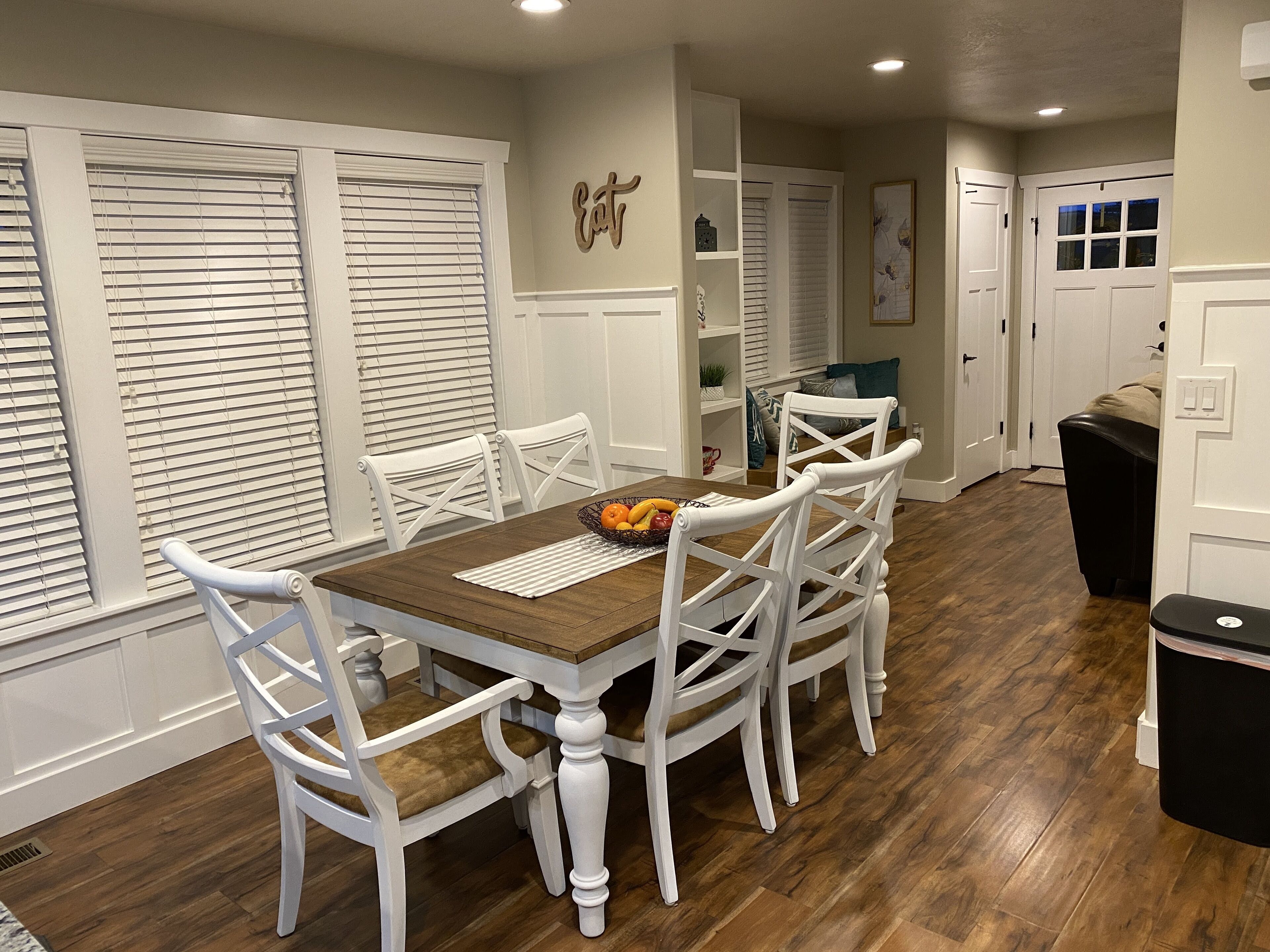 Un comedor con una mesa de madera y sillas blancas, un frutero sobre la mesa y una puerta blanca al fondo.