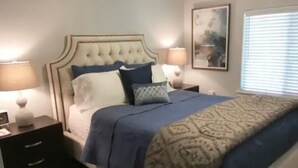 A bedroom with a tufted headboard, a bed with blue bedding, two bedside tables with lamps, and a framed picture on the wall. - Perfect Oasis. NEW Home Away from Home with COVERED PARKING-Close to everything (Stillwater)