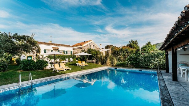 A swimming pool with lounge chairs and umbrellas, surrounded by a well-maintained garden and a stone building with a tiled roof.