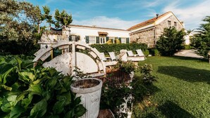 Un bâtiment en pierre avec un toit en tuiles, entouré d’un jardin bien entretenu et d’une grille en métal décorative.