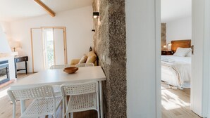Une chambre moderne avec une table blanche, des chaises et un mur en pierre rustique. On aperçoit un lit avec du linge de lit blanc à l’arrière-plan.