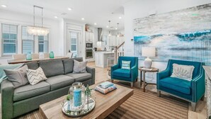 Een moderne woonkamer met een grijze bank, twee blauwe fauteuils, een salontafel, en een keuken op de achtergrond.