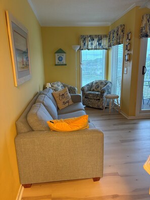 Un salon avec un canapé, un fauteuil, une petite table, une lampe et une fenêtre avec des rideaux.