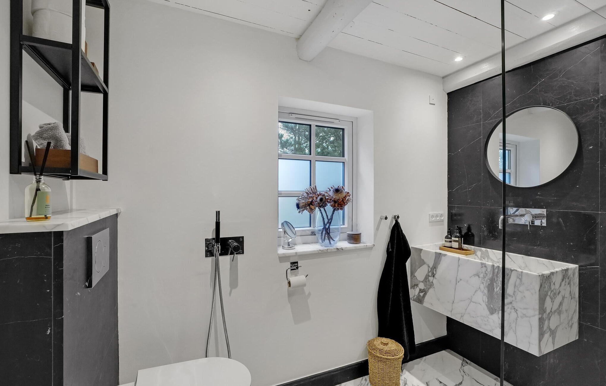 A modern bathroom with a large window, a round mirror, and a marble sink.
