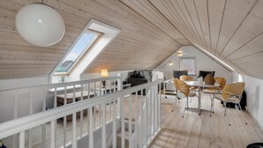 A loft-style room with a high ceiling, wooden beams, a dining area with chairs and a table, a white railing, and a large window. - 3 bedroom gorgeous home in Vestervig (Vestervig)