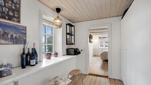 A narrow hallway with a wooden ceiling, a window with a view of the outside, a small table with a floral cushion, and a shelf with bottles and a box. - 3 bedroom gorgeous home in Vestervig (Vestervig)