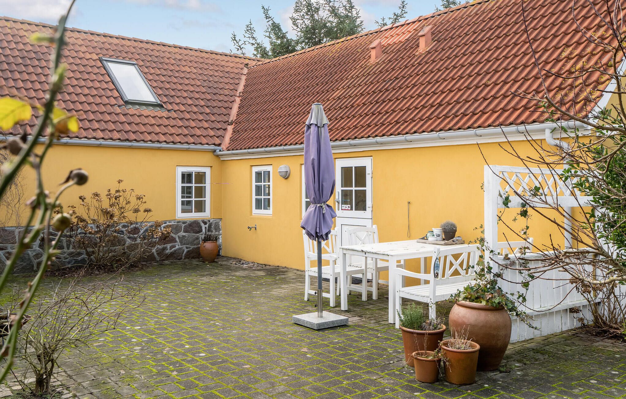 Une cour avec un sol carrelé, une table et des chaises blanches, un parasol violet et des plantes en pot.
