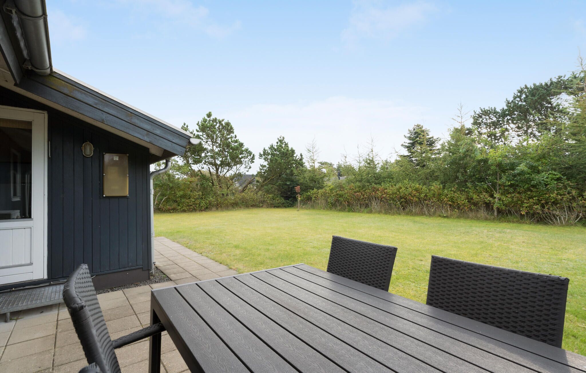 A patio with a table and chairs, a small building with a door and window, and a grassy yard with trees.