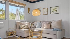 A cozy living room with a sofa, a round table, and a wicker chair. There are framed botanical prints on the wall and a hanging light fixture. - 3 bedroom awesome home in Skjern (Skjern)