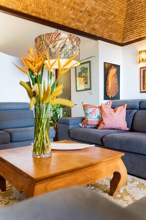 A living room with a blue sofa, a wooden coffee table, and a vase with yellow flowers. - Magnificent 5 Br Villa with pool: amazing views (Puna'auia)