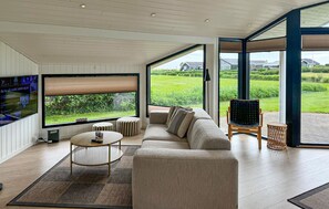 A modern living room with a large TV, comfortable seating, and a view of a green landscape. - Amazing home in Sæby with sauna (Sæby)