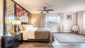 A modern hotel room with a large bed, a ceiling fan, a view of the desert landscape, and a leather armchair. - Onsen Hotel & Spa (Desert Hot Springs)