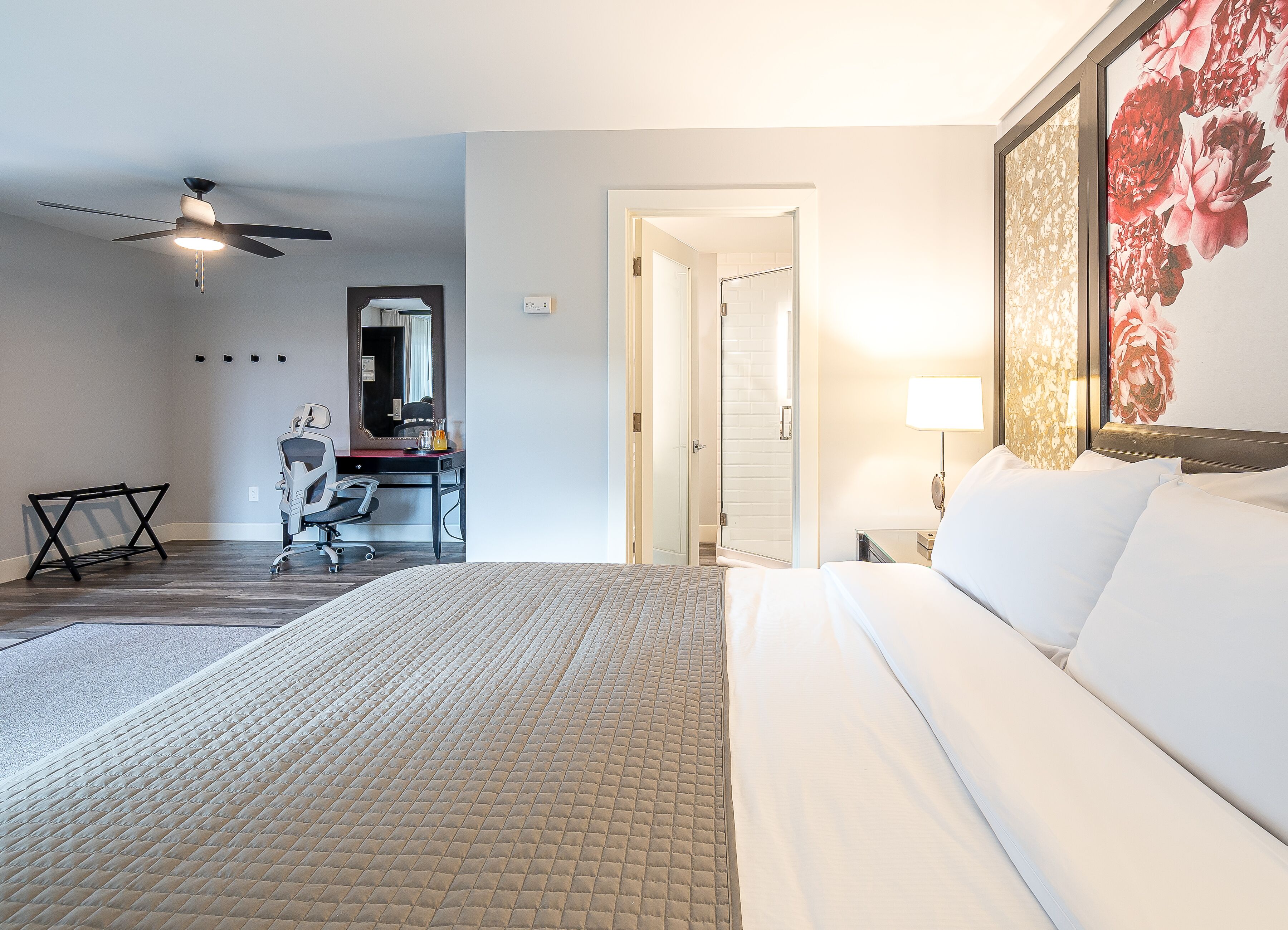 A hotel room with a bed, a desk with a chair, a ceiling fan, and a bathroom door.