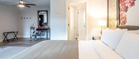 A hotel room with a bed, a desk with a chair, a ceiling fan, and a bathroom door.