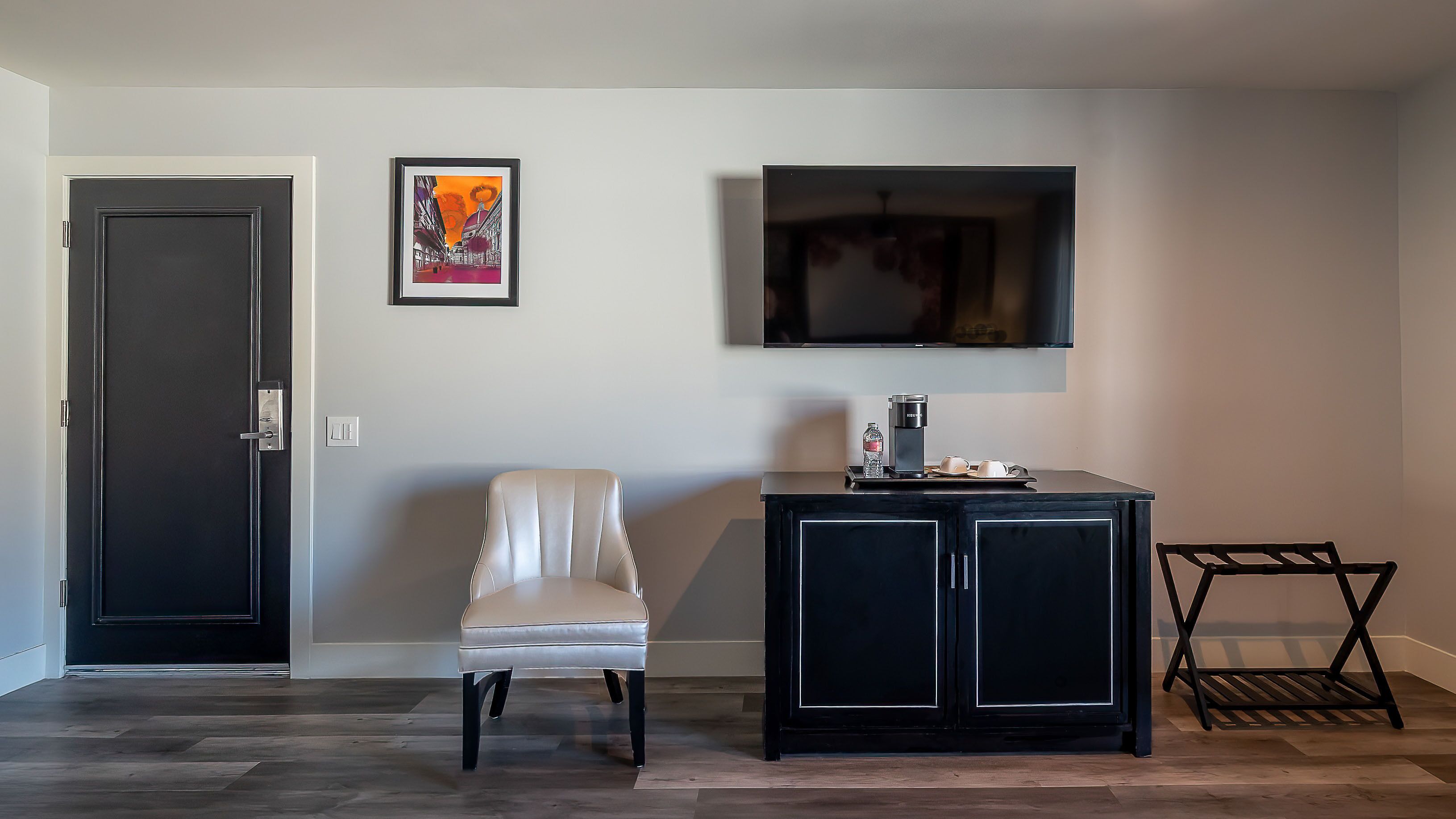 A room with a door, a framed artwork, a television, a chair, a coffee station, and a small table.