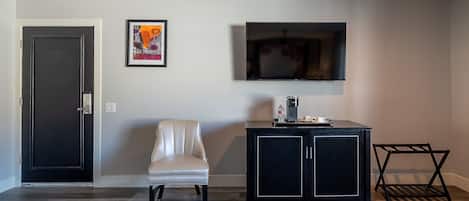 A room with a door, a framed artwork, a television, a chair, a coffee station, and a small table.