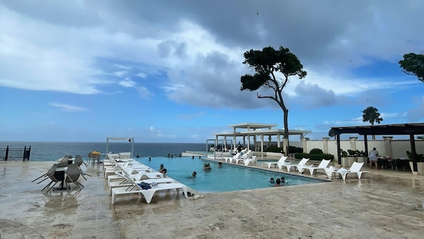A pool area with lounge chairs and a covered seating area, overlooking the sea.