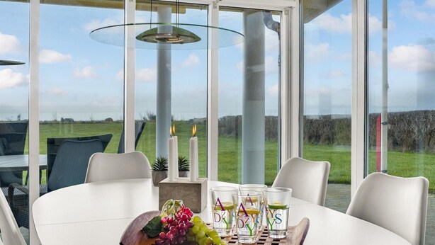 A modern dining area with a table set for four, featuring a glass centerpiece with fruits and glasses, and a view of a grassy field through large windows. - 3 bedroom amazing home in Spøttrup (Spøttrup)