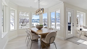 A modern kitchen with a wooden table, four chairs, a hanging light fixture, and large windows offering a view of a snowy landscape.