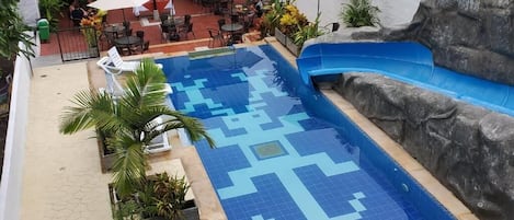 Una piscina con un diseño geométrico de azulejos, rodeada de sillones de playa y sombrillas, y una pared rocosa en un lateral.