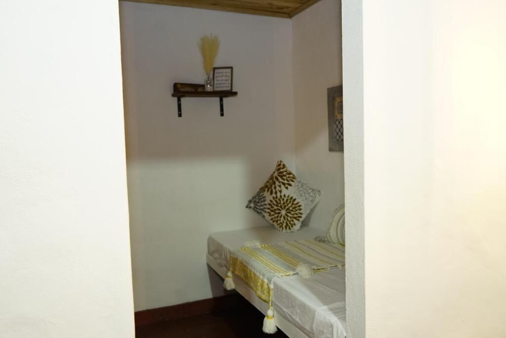 A small bedroom with a single bed, a nightstand, a wall-mounted shelf with a vase and a framed picture, and a decorative pillow.