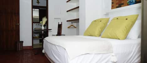 Un dormitorio con puerta de madera, cama con ropa de cama blanca y almohadas amarillas, una mesita de noche pequeña y un espejo.