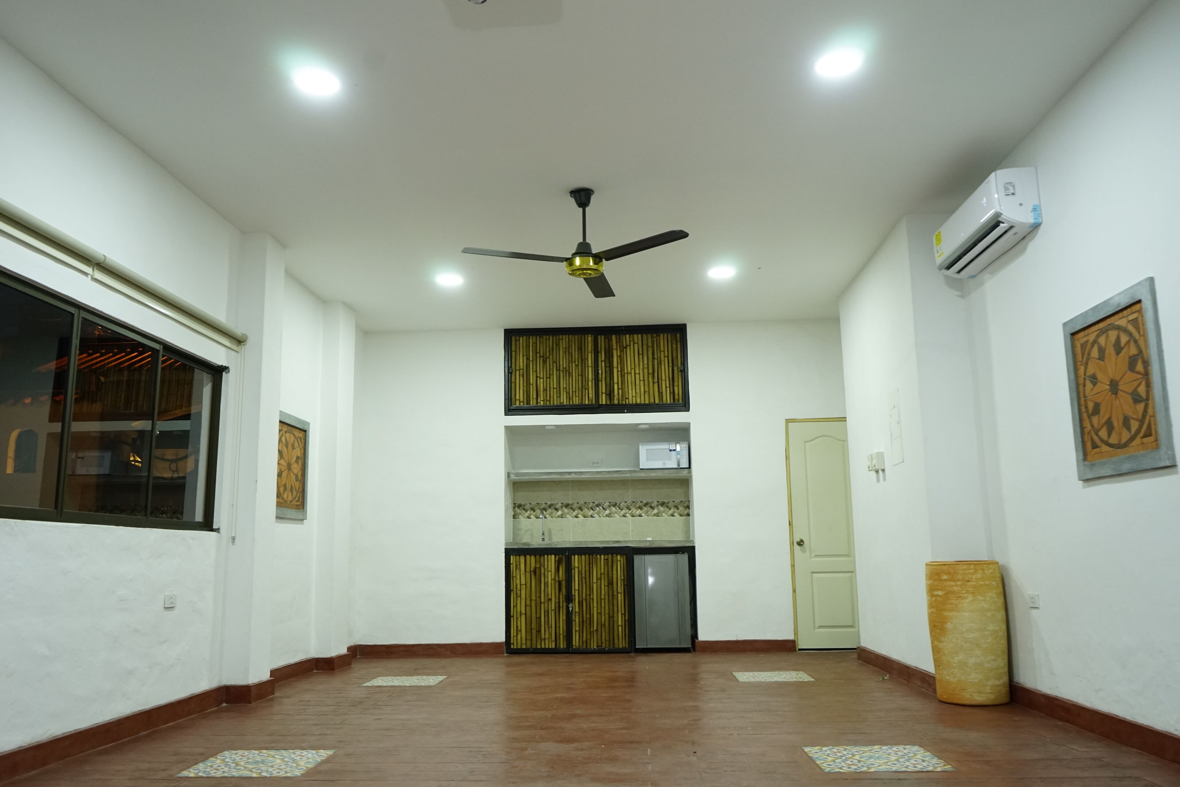 A spacious room with a ceiling fan, an air conditioning unit, a window with blinds, a door, and a decorative wall piece.