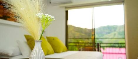 Quarto com uma cama, um vaso branco com grama seca e flores brancas, e vista para colinas verdes através de uma porta deslizante.