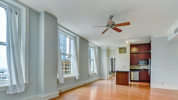 A spacious room with hardwood flooring, large windows with white curtains, a ceiling fan, and a kitchen area with wooden cabinets and appliances. - Popular Filming Area- Downtown Atlanta Condo (atlanta)