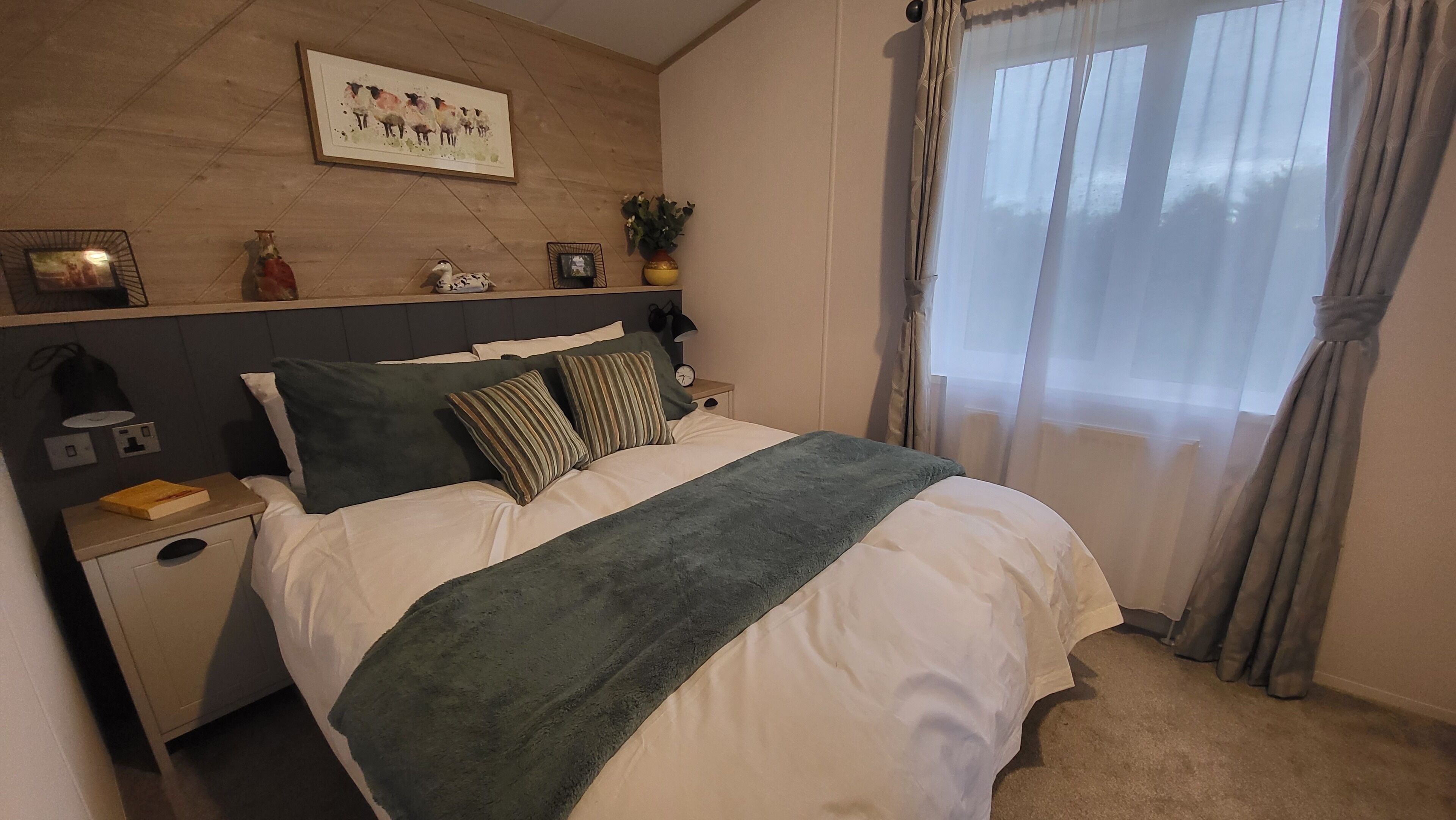 A neatly arranged bedroom with a bed, bedside tables, a nightstand, a wall-mounted shelf with decorative items, and a window with curtains.
