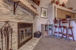 A living room with a stone fireplace, wooden chairs, a bookshelf, and a kitchen area with wooden cabinets. - Winter Loft Retreat with Jacuzzi, Pool & Fireplace (Brian Head)