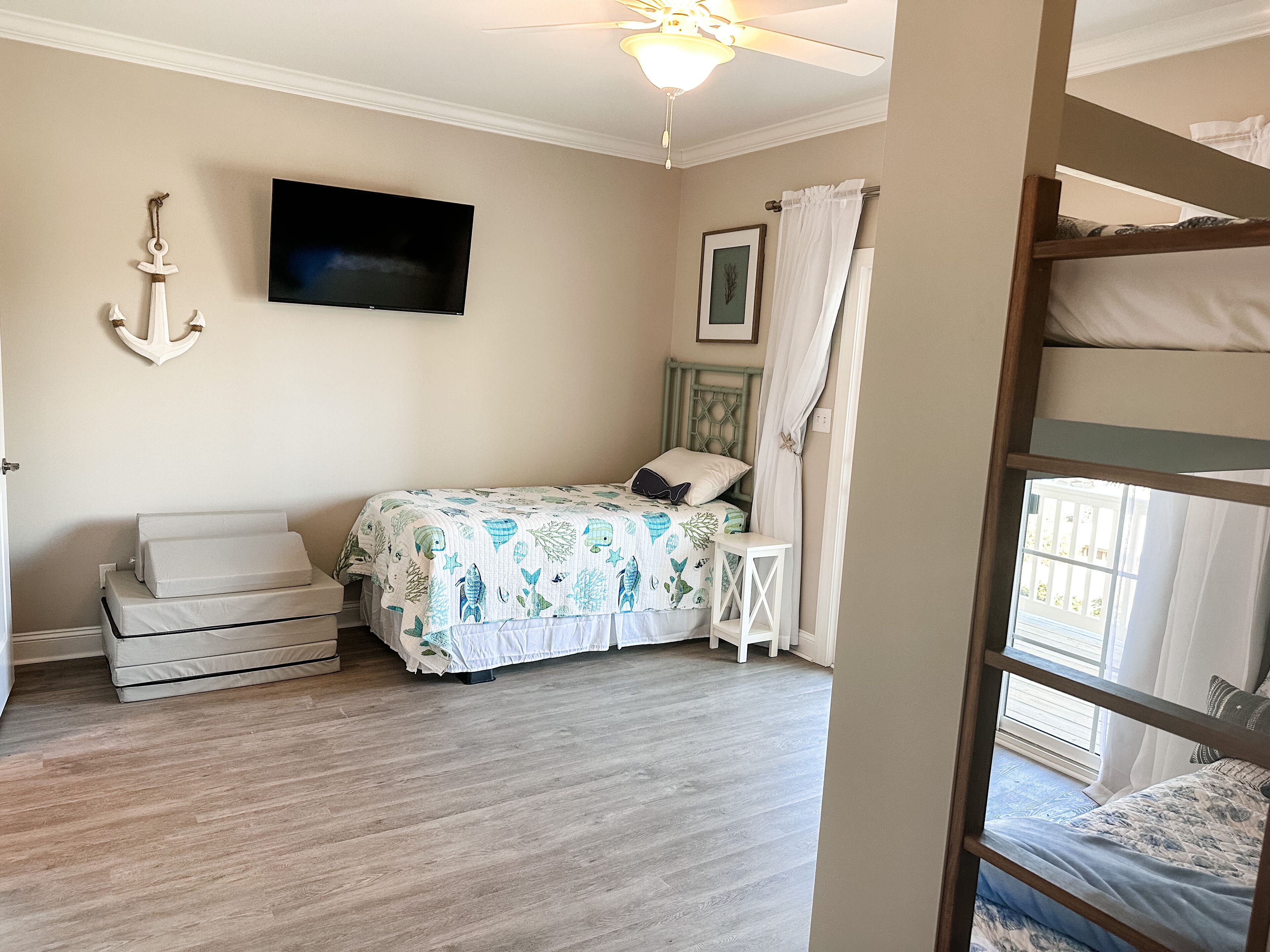 A bedroom with a bunk bed, a single bed with a patterned bedspread, a television on the wall, and a ceiling fan.