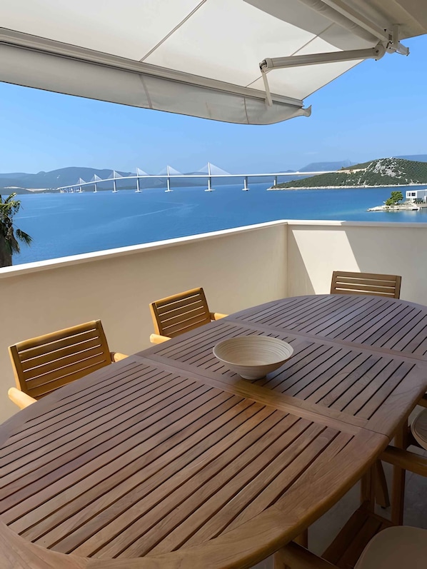 A wooden table with a bowl, surrounded by chairs, overlooking a body of water with a bridge in the distance.