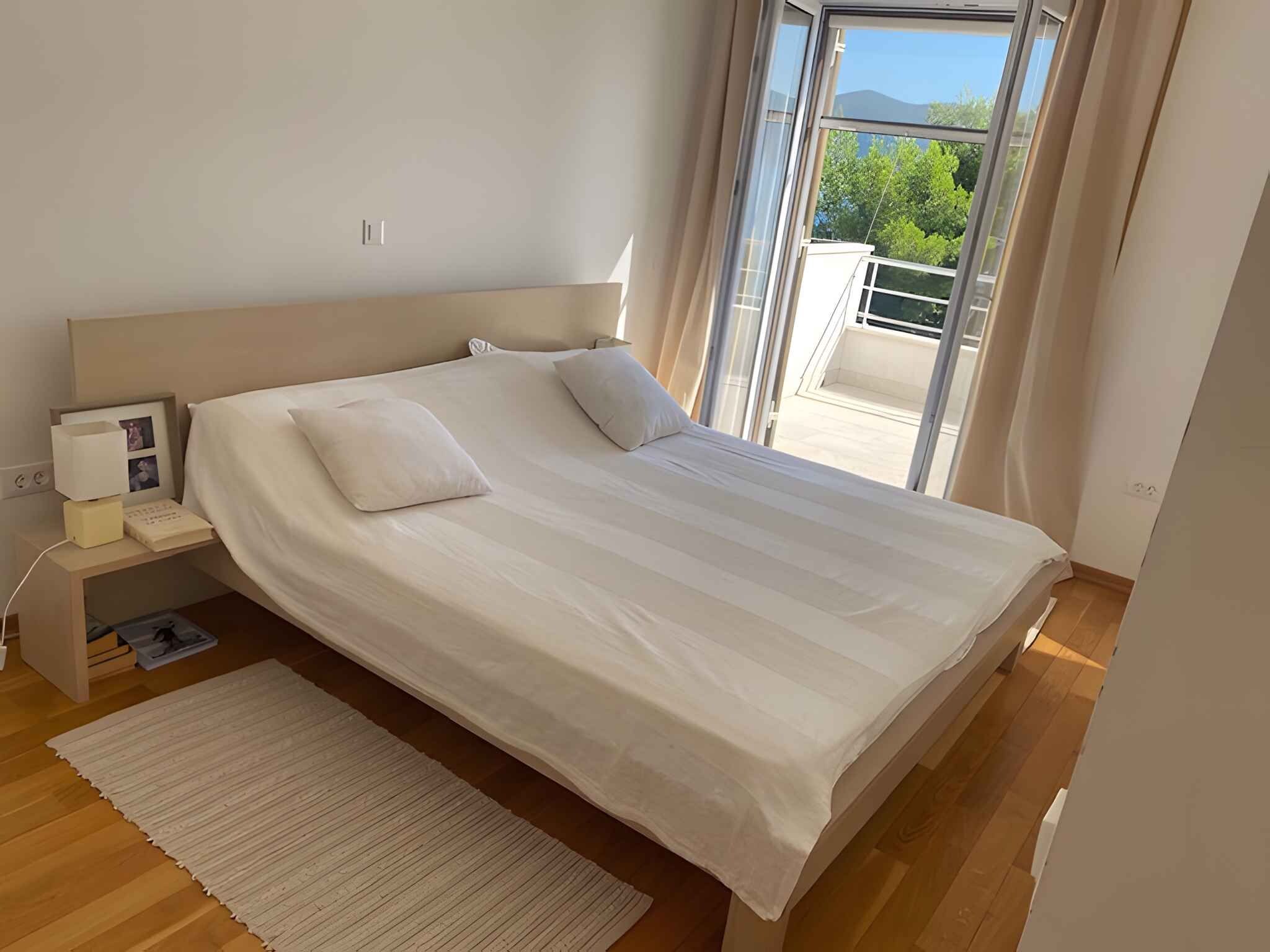 A modern bedroom with a large bed, a bedside table with a lamp, and a view of greenery through a glass door.