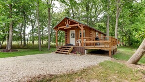 Eine Blockhütte mit Veranda, einer Terrasse und einer Zuwegung mit Kies, umgeben von Bäumen.