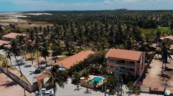 An aerial view of a resort with multiple buildings, a swimming pool, and dense palm trees.