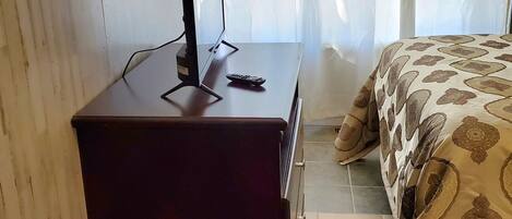 A hotel room with a wooden desk, a television, a remote control, a bed with patterned bedding, and curtains.