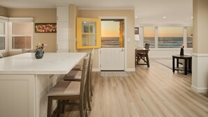 Een ruime keuken met een groot eiland, houten stoelen en uitzicht op zee door een geopende deur.