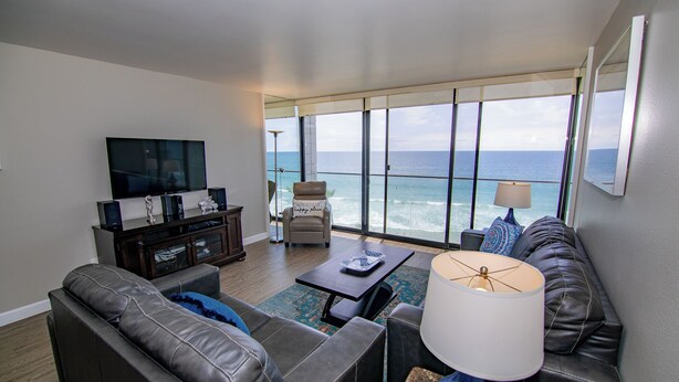 A living room with a large window overlooking the ocean, a flat-screen TV, leather sofas, and a coffee table. - ONE BEDROOM CONDO 402 FULL BEACH AND OCEAN VIEW (San Diego)