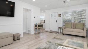 Un salon avec un canapé beige, un téléviseur fixé au mur, un ventilateur de plafond et une fenêtre avec des stores.