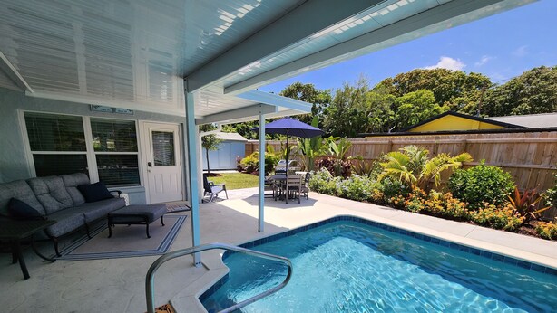 Une piscine dotée d’une balustrade en verre, un patio couvert aménagé avec du mobilier d’extérieur, et une vue sur un jardin avec une clôture en bois et de la verdure.