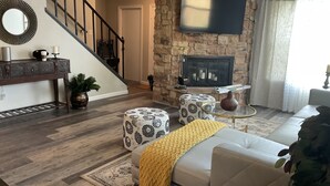 A living room with a stone fireplace, a flat-screen TV mounted above it, a staircase, and a sofa with a yellow throw blanket. - Entire House - 5 Minutes Drive to AT&T Stadium (Arlington)