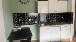 Een kleine keuken met een zwart aanrecht, witte kasten, een zwart-wit marmeren spatwand, een wandklok en een magnetron.