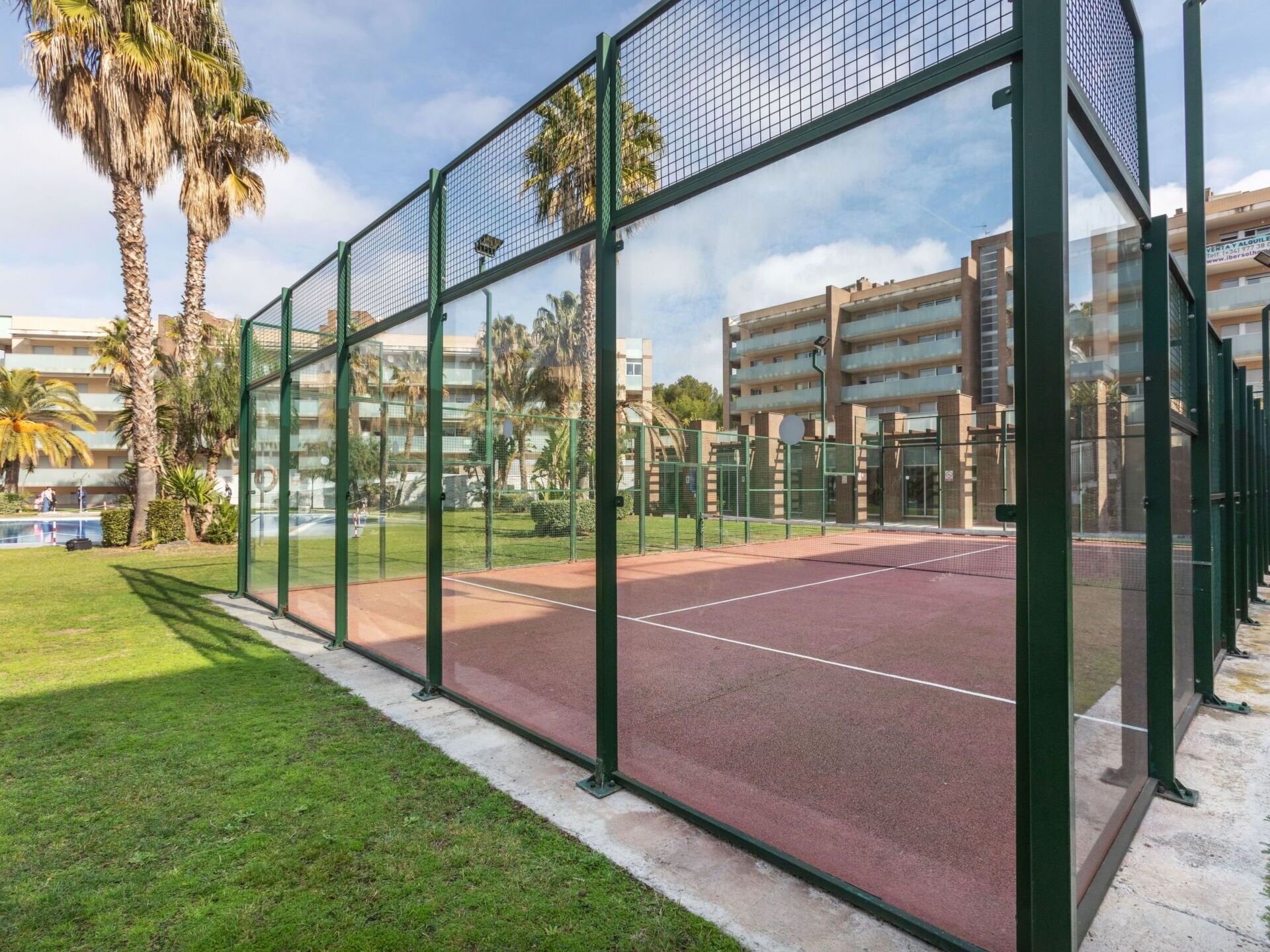 Una cancha de tenis rodeada por una cerca metálica verde, con una vista despejada de un edificio residencial al fondo.