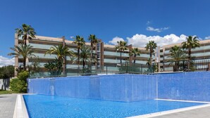 Une piscine dotée d’une terrasse carrelée, entourée de palmiers et bordée d’un bâtiment moderne avec de nombreux balcons.