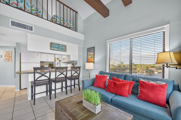 A living room with a blue sofa, red cushions, a coffee table with a plant, a dining area with a table and chairs, and a kitchen with white cabinets.