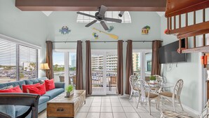 A spacious living area with a ceiling fan, a balcony with curtains, a dining set, and a TV mounted on the wall.