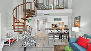A spacious living area with a wooden spiral staircase, a kitchen with a central island, and a dining area with a blue sofa and red cushions.