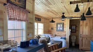 A cozy cabin interior with a wooden ceiling, a bed with a floral bedspread, a dining table with chairs, and a kitchen area with a radio. - Brand new tiny cabin located on farm. Near rivers, trailsheads & national forest (West Plains)