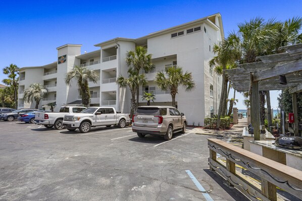 A parking lot with several SUVs and a white truck, a building with multiple balconies and palm trees, and a wooden pergola with outdoor seating. - Corner Beachfront Condo at Cabana Club 201 w/ Panoramic Gulf Views & Private Beach Access (Destin)